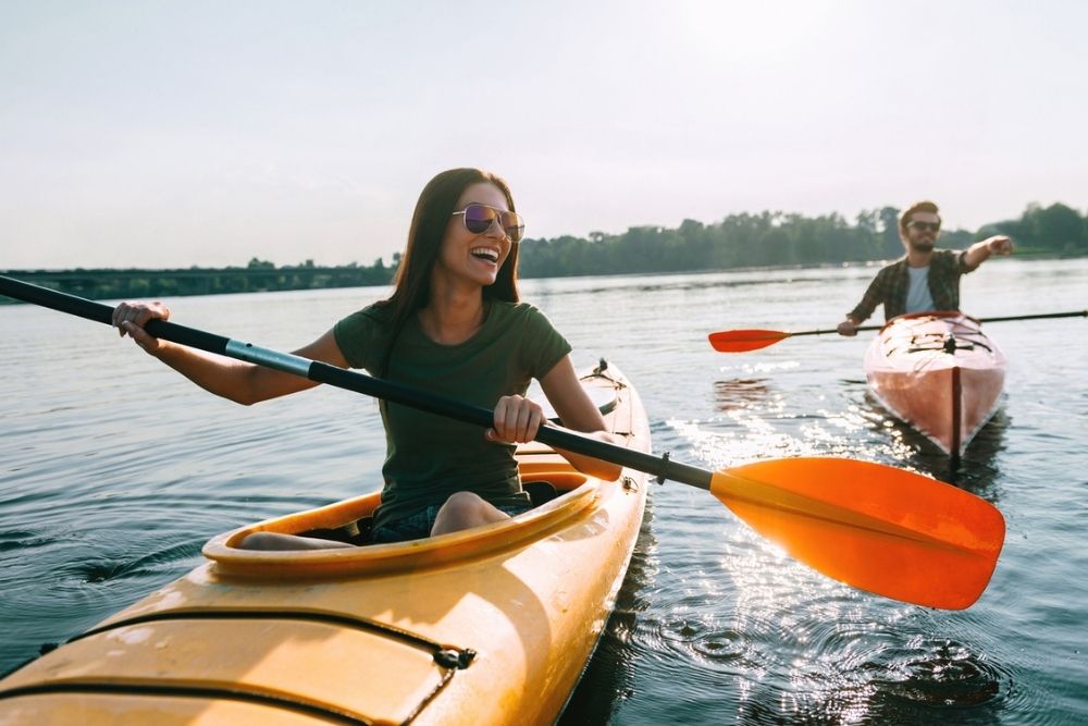 Two people kayaking on a calm lake, both smiling and wearing sunglasses. The sun is shining, and trees line the distant shore.
