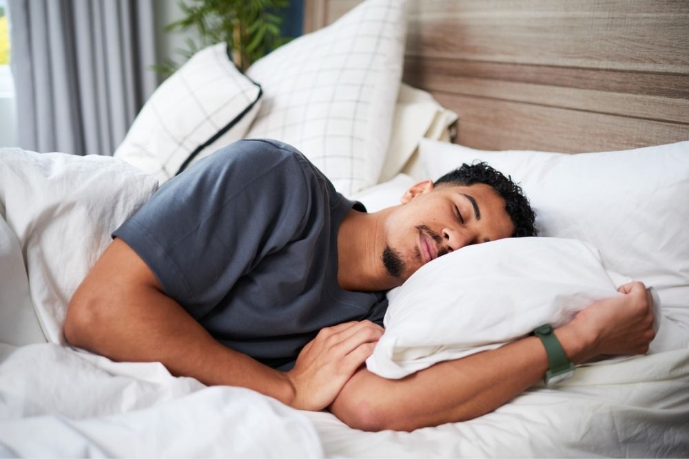 A man with short hair is sleeping peacefully on his side in a bed with white sheets and pillows, wearing a gray shirt and a green wristband.