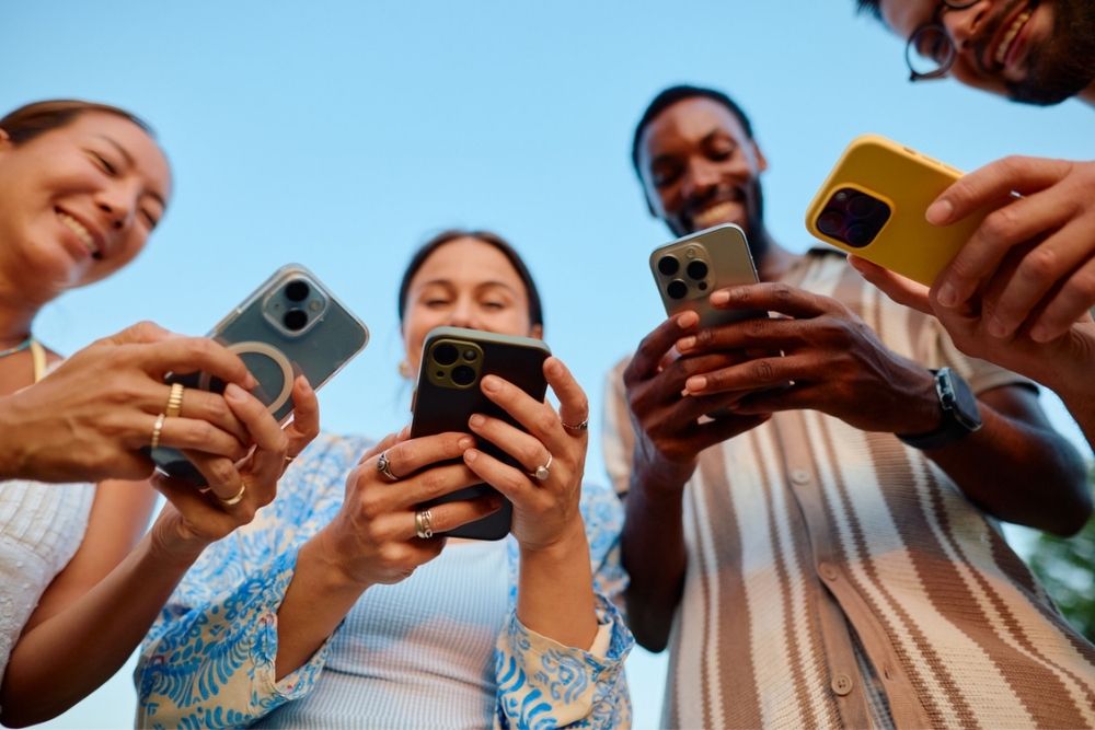 Four people stand in a circle outdoors, smiling and looking down at their smartphones, holding them out in front of themselves against a clear blue sky.