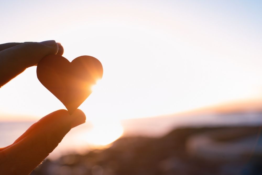 A hand holds a small heart-shaped object against the sunset, with sunlight shining through the heart and a blurred beach in the background.