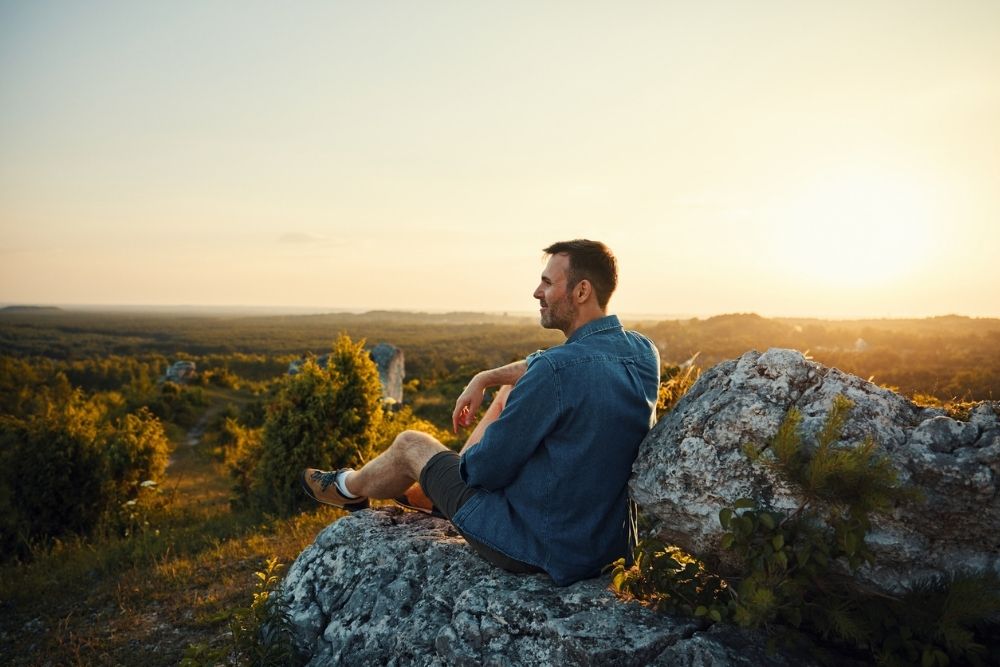 A man sits on a large rock, gazing at the scenic landscape during sunset. The sky is golden, and the area is surrounded by greenery and rolling hills.