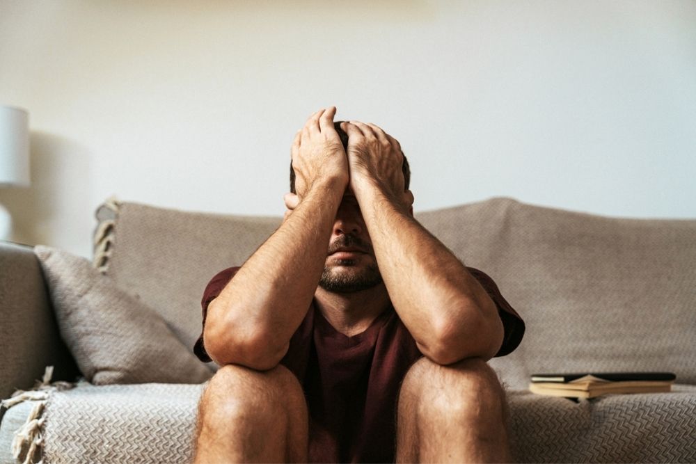 A man sits on the floor in front of a couch, covering his face with both hands. An open book lies beside him, suggesting he may be feeling stressed or overwhelmed.
