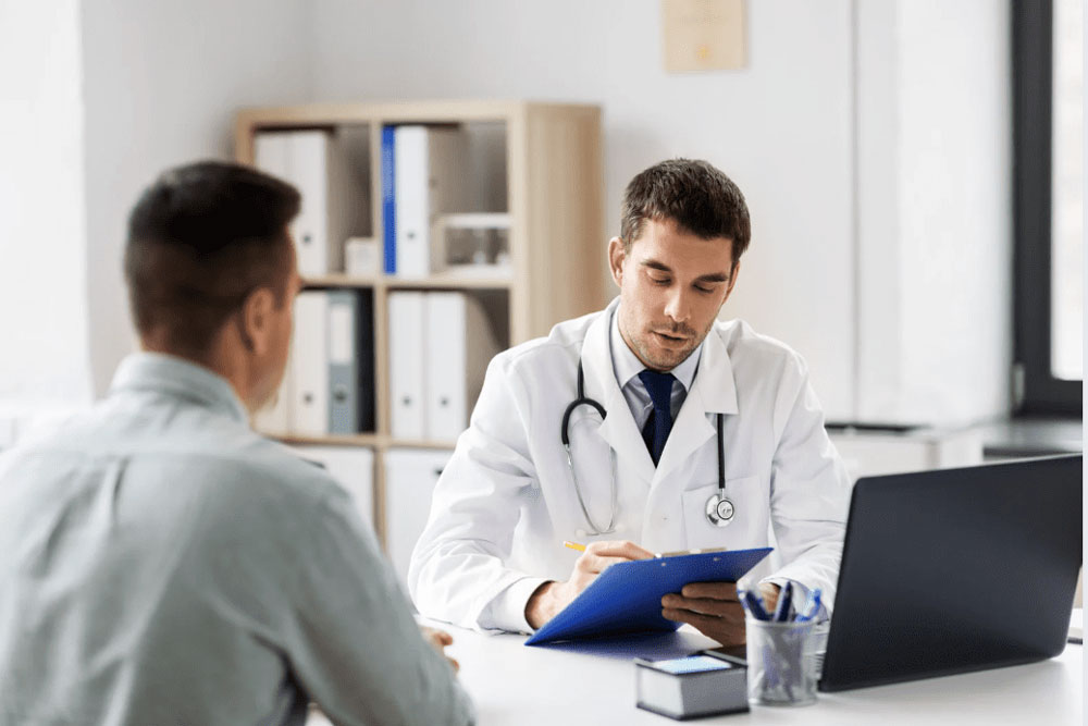 A doctor in a white coat, holding a clipboard, listens and takes notes while sitting across from a patient in an office with a laptop and shelves in the background.