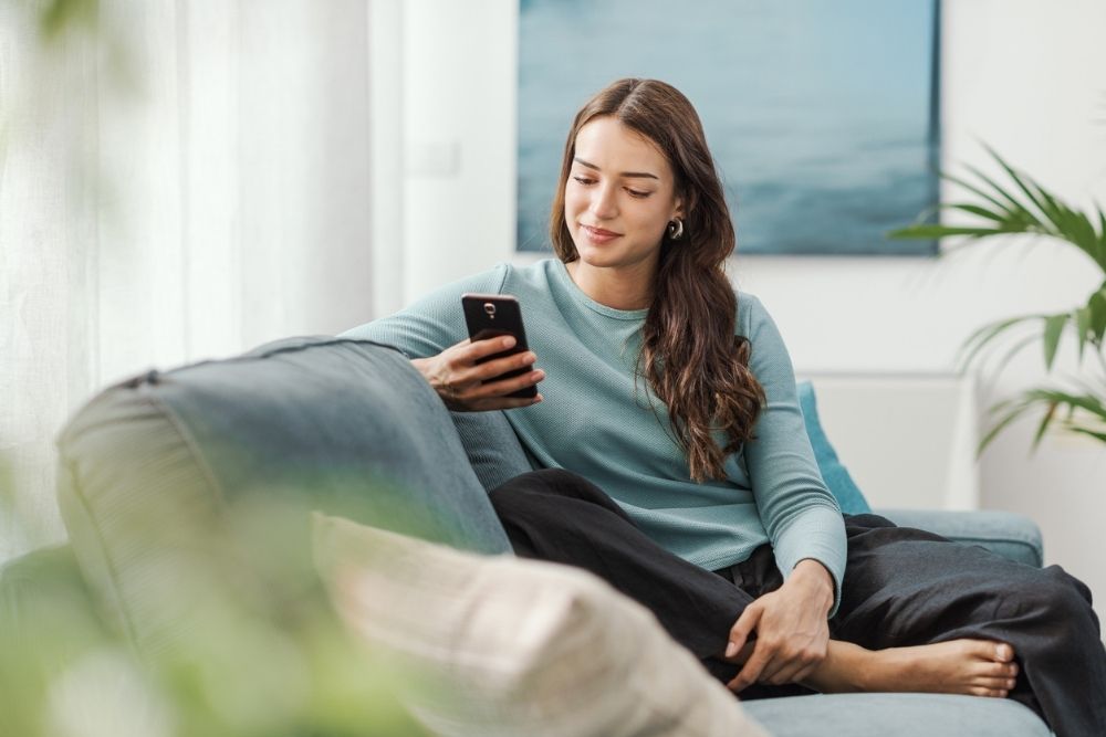 A woman sits comfortably on a sofa, smiling as she looks at her smartphone. She is dressed casually in a light blue top and dark pants, with natural light coming through a nearby window.