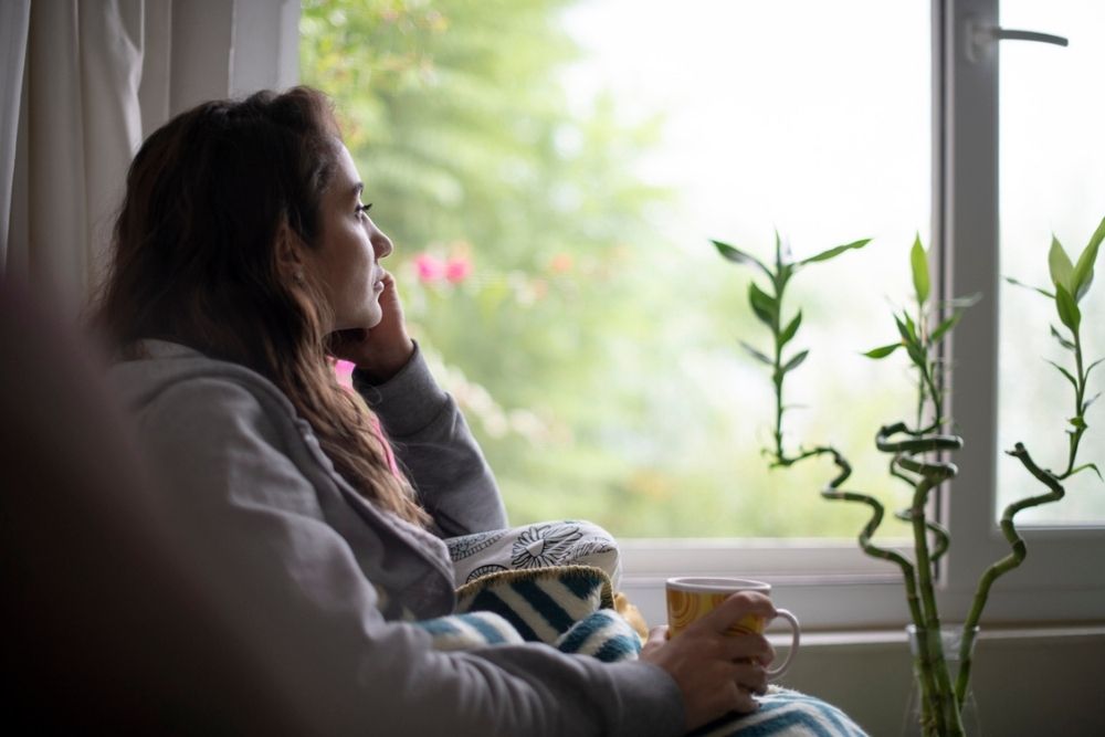 A woman sits by a window, wrapped in a blanket and holding a mug, gazing outside thoughtfully. Green plants and soft natural light are visible near her.