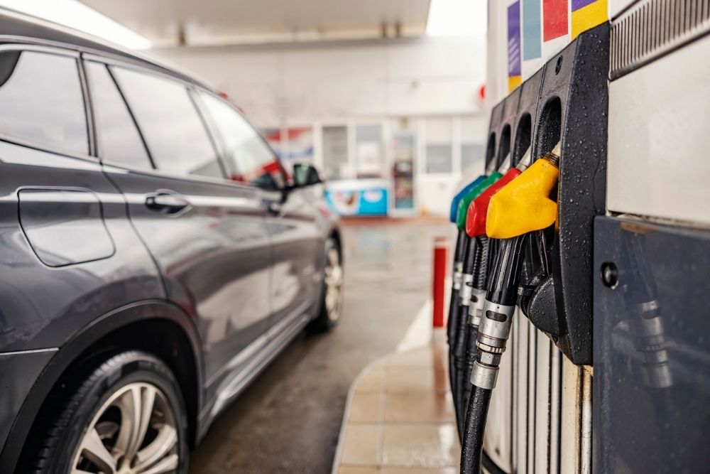 A gray car parked at a gas station next to a fuel pump with multiple colorful nozzles. The scene is slightly wet, suggesting recent rain.