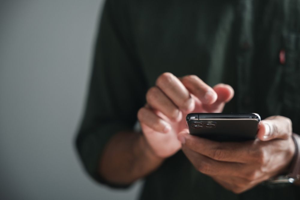 A person in a dark green shirt uses a smartphone, tapping the screen with one hand and holding the device with the other, against a neutral background.