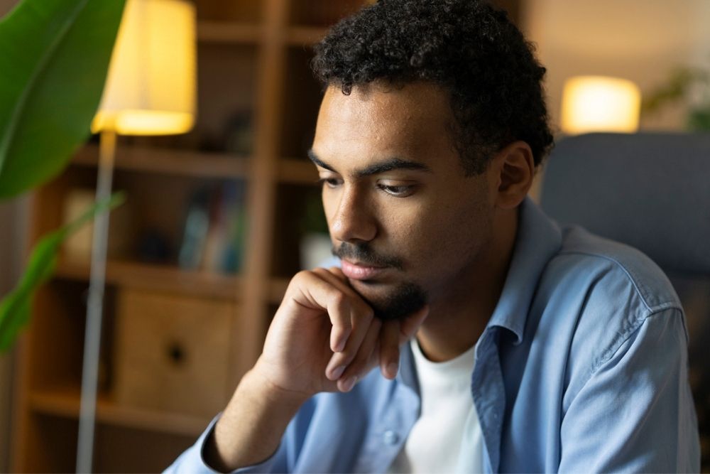 A man with short curly hair, a beard, and a blue shirt sits indoors with his hand on his chin, looking down thoughtfully. Warm lighting and blurred shelves are visible in the background.