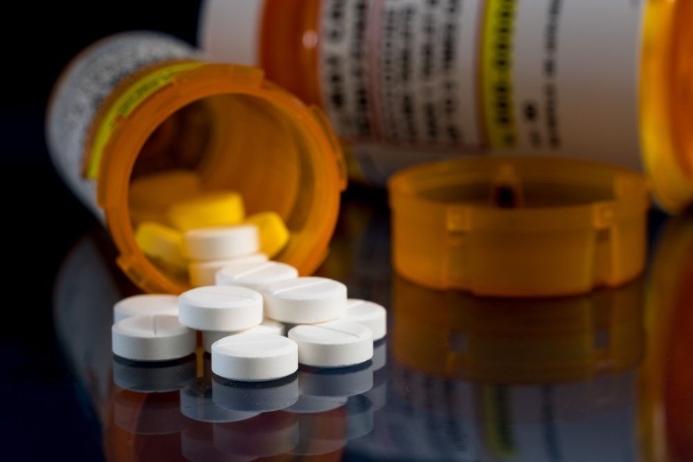 White tablets spilling from an orange prescription bottle, with another bottle and lid in the background, all on a dark reflective surface.