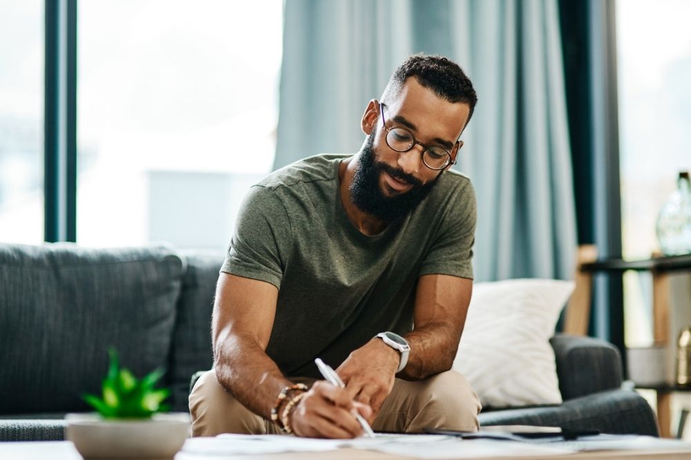A man with glasses and a beard sits on a couch, smiling as he writes on paper at a coffee table in a bright, modern living room.
