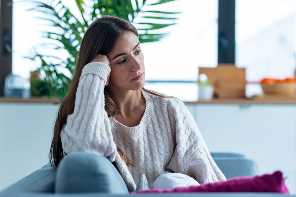 A woman in a white sweater sits on a couch, resting her head on her hand and looking thoughtful or concerned, with a plant and kitchen in the background.