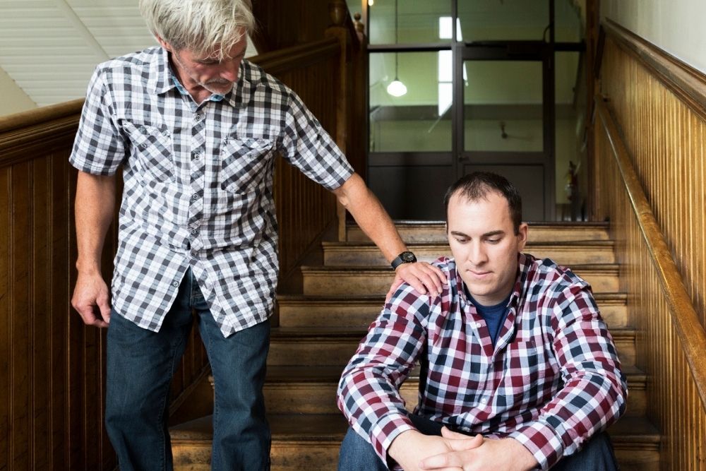 An older man stands on stairs, comforting a younger seated man with his hand on his shoulder. Both wear plaid shirts; the younger man looks down, appearing upset.