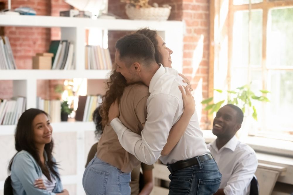 Two people hug in an office while three colleagues smile and watch. Everyone appears happy, and bookshelves and windows are visible in the background.