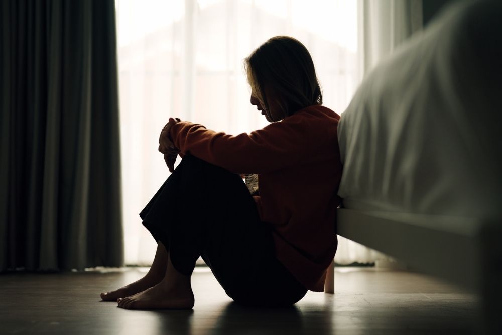 A person sits on the floor beside a bed, hugging their knees, silhouetted against a window with sheer curtains, appearing thoughtful or sad.