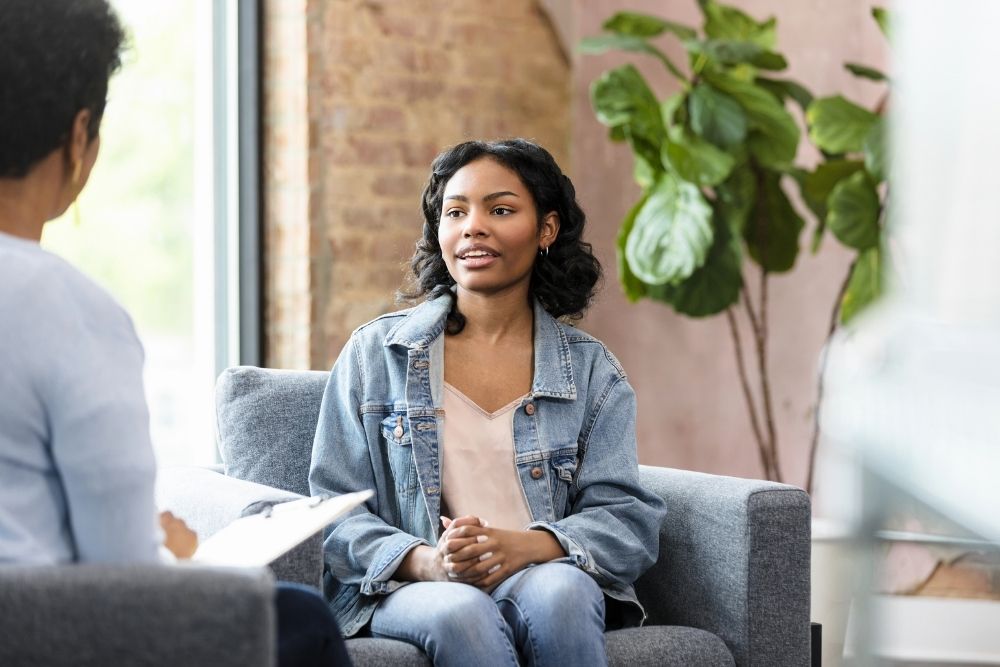 A young woman in a denim jacket sits on a gray couch, talking to another person whose back is to the camera. There is a large green plant in the background and natural light coming in from a window.