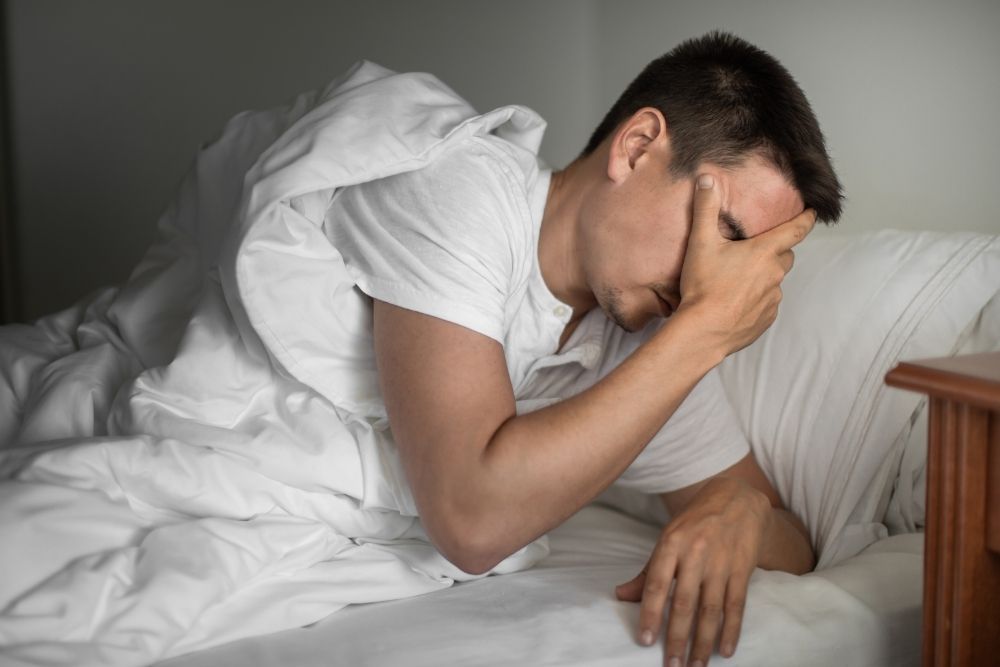 A man sits up in bed holding his head with one hand, appearing to be in pain or distress. He is covered with a white comforter and wears a white t-shirt.