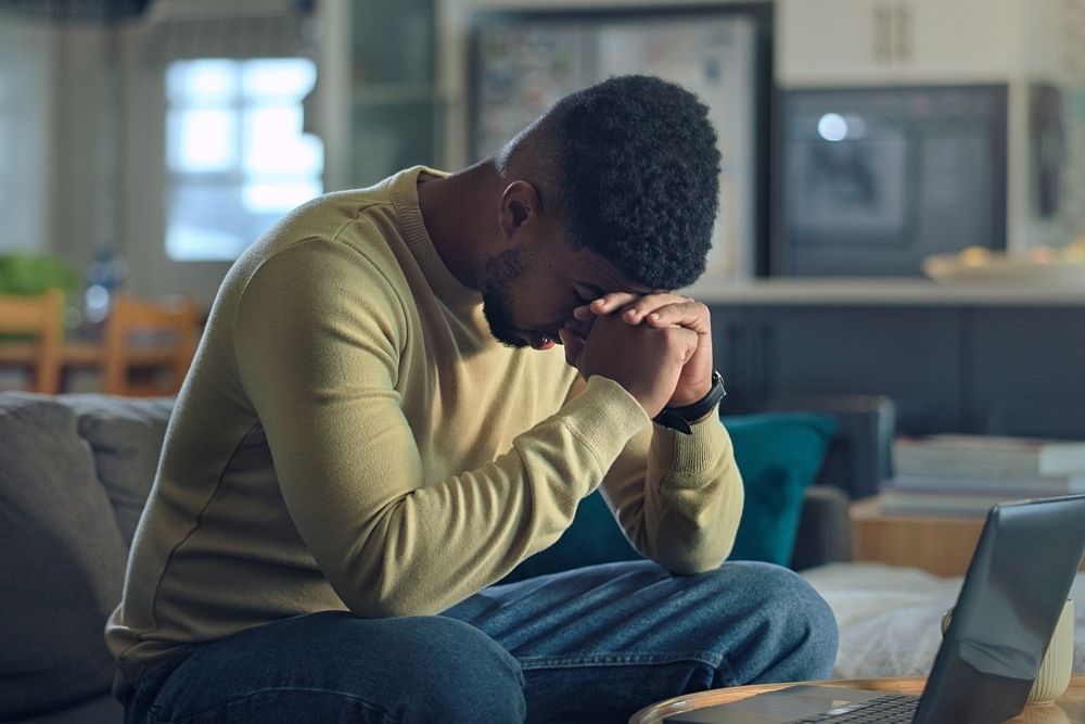 A man sits on a couch with his elbows on his knees and hands clasped, head bowed, looking stressed or deep in thought in front of an open laptop.