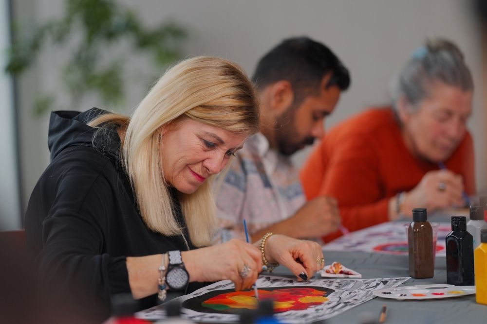 Three adults sit at a table, focused on painting colorful artwork. The woman in the foreground smiles as she paints, while the others work intently in the background. Art supplies are visible on the table.