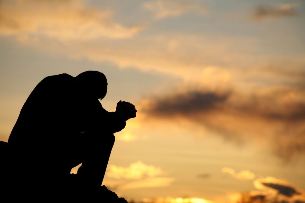 A person sits with head bowed and hands clasped, silhouetted against a dramatic sunset sky with clouds.
