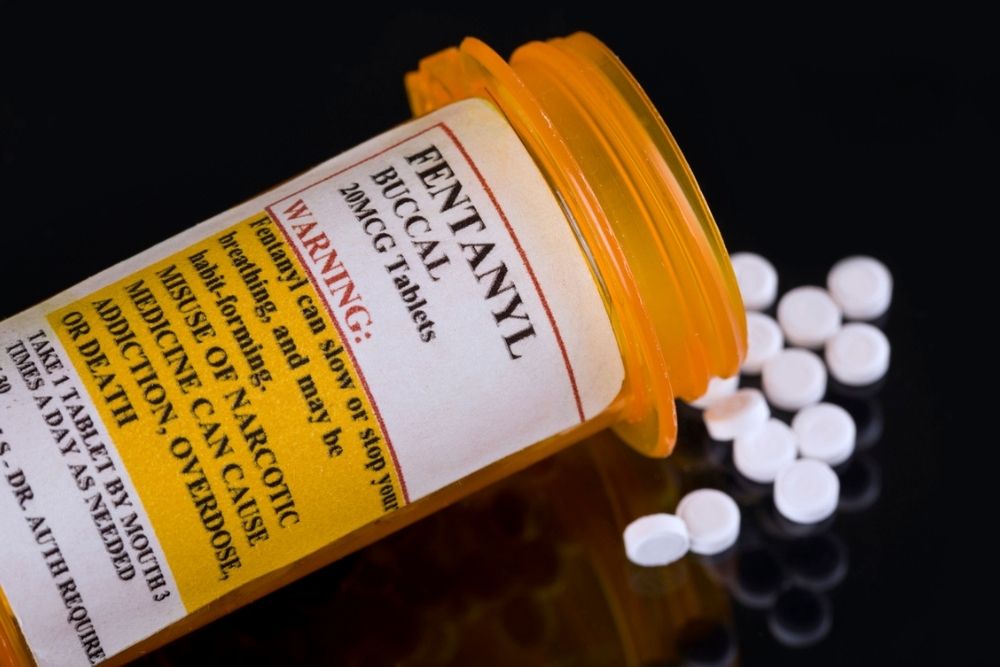 An orange prescription bottle labeled Fentanyl lies on its side with white tablets spilling out. The label includes a warning about addiction and overdose risks. The background is black.