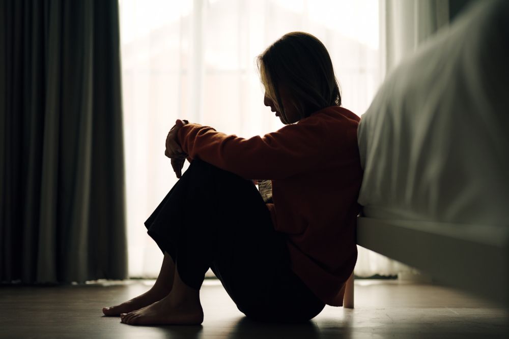 A person sits on the floor beside a bed, hugging their knees and looking down, silhouetted by soft light coming through a window with curtains.