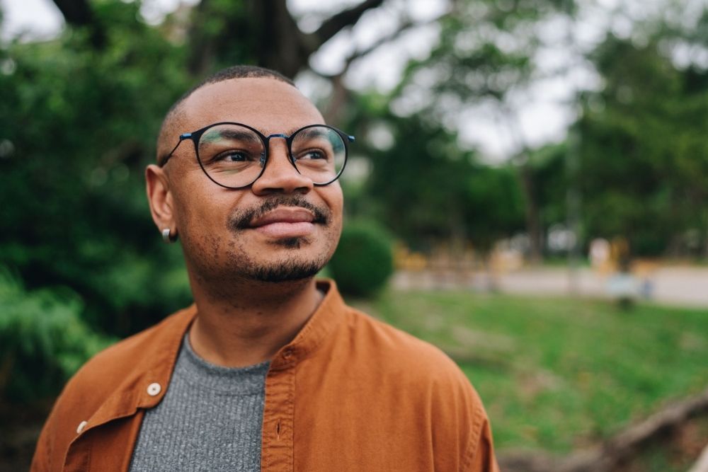 A person wearing glasses and a brown jacket stands outdoors, looking up and smiling slightly. The background is blurred greenery and trees.