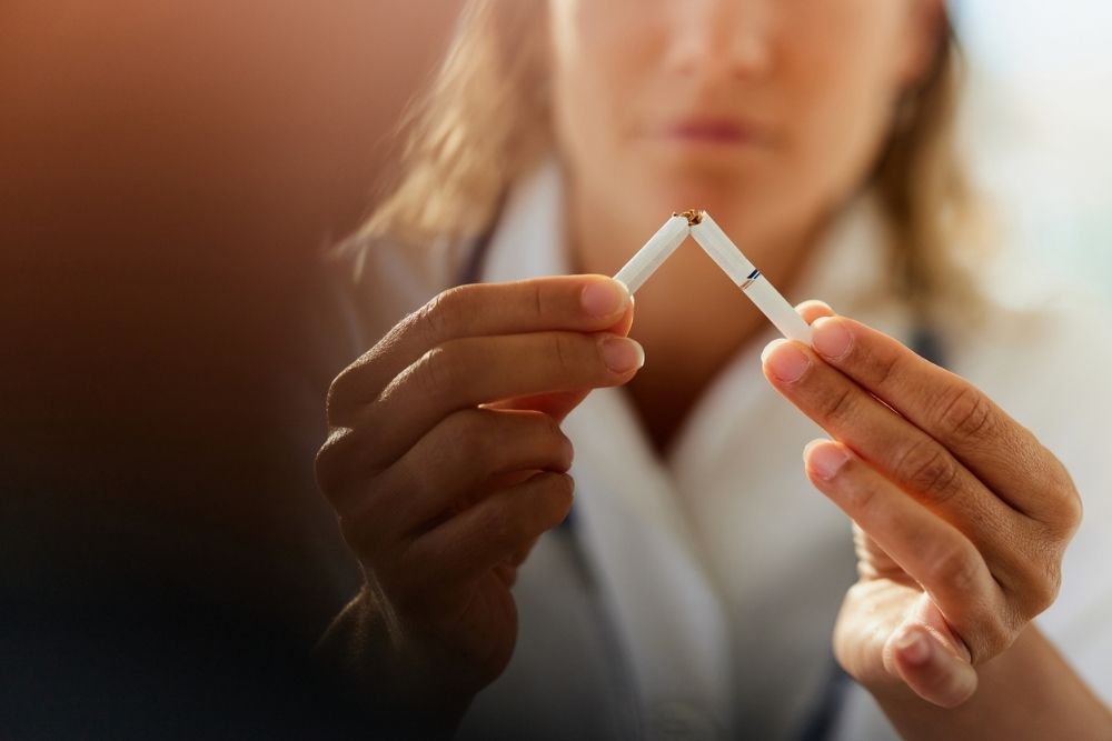 A person holds a broken cigarette in both hands, symbolizing quitting smoking. The background is blurred, focusing on the cigarette and the persons hands.