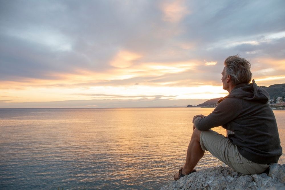 A person sits on a rocky shore, looking out over a calm sea at sunset. The sky is filled with soft clouds and warm colors, while distant hills line the horizon.