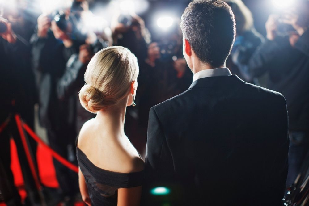 A woman in an elegant black dress and a man in a suit stand on a red carpet facing photographers, with bright camera flashes lighting up the scene.