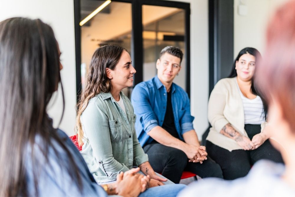 Five people sit in a circle indoors, engaged in conversation. They appear to be in a casual meeting or support group, with some smiling and listening attentively. The setting is modern with glass doors in the background.