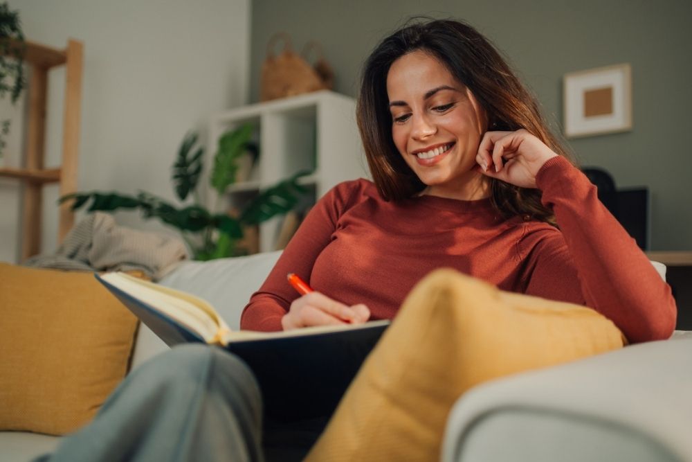 A woman sits on a couch, smiling as she writes in a notebook with an orange pen. She is relaxed, surrounded by pillows, with indoor plants and shelves in the background.