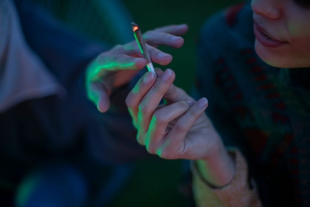 Two people pass a lit joint between them, illuminated by dim greenish light. Only their hands and part of their faces are visible, with an out-of-focus background.