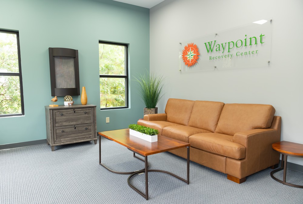 A waiting room with a tan sofa, wooden coffee table with a plant, dresser with decor, two windows, and a Waypoint Recovery Center sign on a light blue wall.