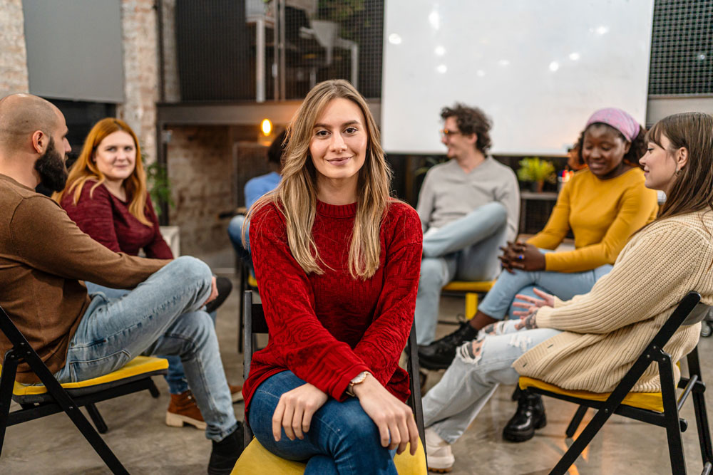 A woman in a red sweater sits facing the camera, smiling, while five other people sit in a circle behind her, talking and interacting in a casual indoor setting.