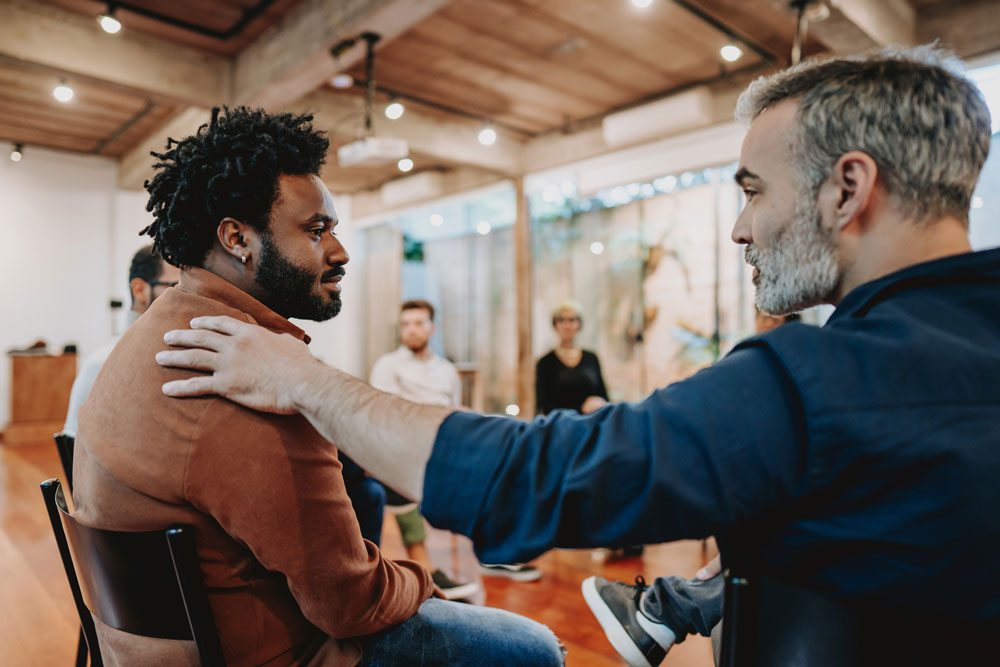 A man comforts another man by placing a hand on his shoulder during a group meeting in a modern, well-lit room as others sit and observe in the background.