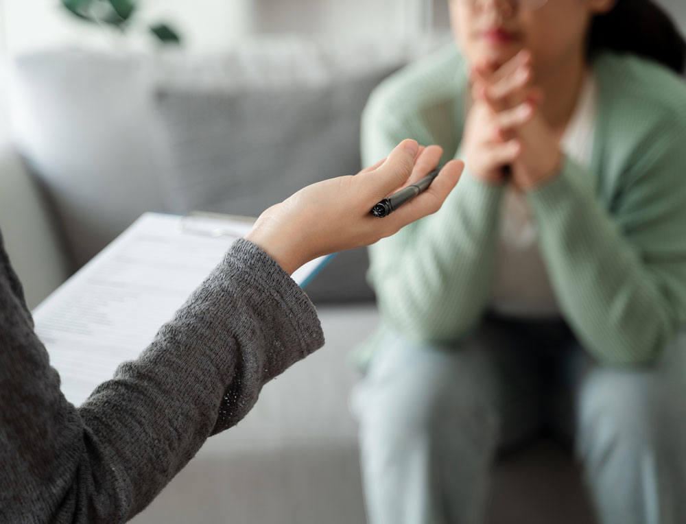A person holding a pen and clipboard speaks to another person who sits with hands clasped under their chin, suggesting a counseling or therapy session in a relaxed indoor setting.