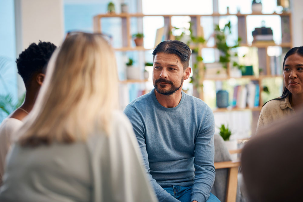 A man in a blue sweater sits in a group discussion with three others in a bright room with shelves and plants in the background.