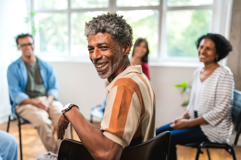 A smiling man sits in a support group circle, looking back over his shoulder. Three other people are seated in the background, all appearing engaged and happy in a bright, sunlit room.