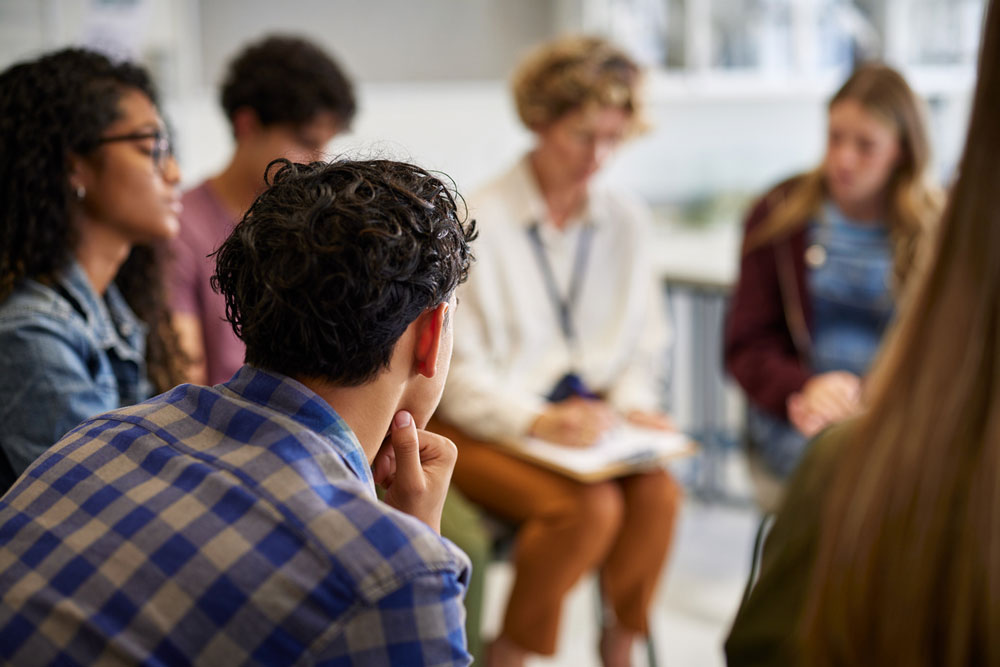 A group of people sit in a circle having a discussion. The focus is on the back of a person with curly hair in the foreground, while others appear blurred in the background.