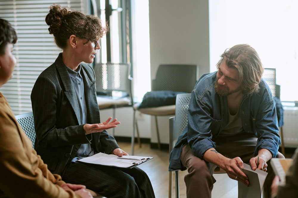 Three people sit in a circle indoors. One woman, holding a clipboard, gestures while talking. A man next to her listens, leaning forward. They appear to be in a group discussion or therapy session.