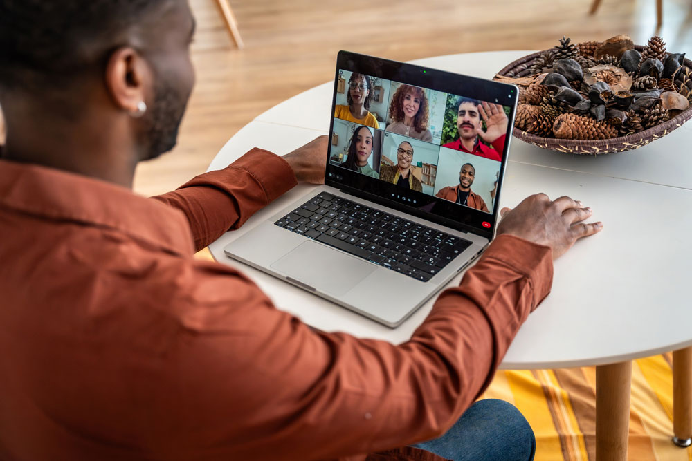 A person sits at a table using a laptop for a video call with six people. A decorative bowl with pinecones is on the table.
