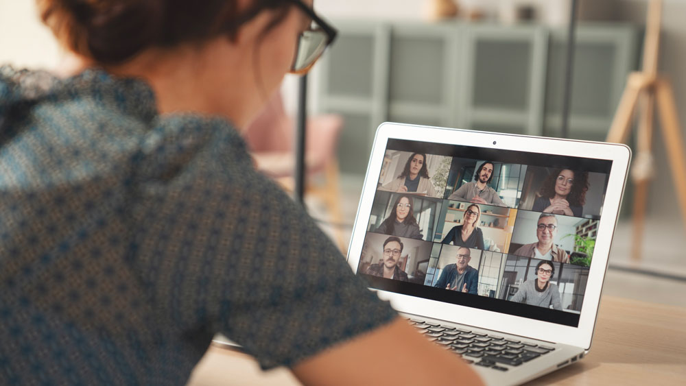A woman sits at a desk, looking at a laptop screen displaying a video call with nine people, each in their own video window, participating in a virtual meeting.