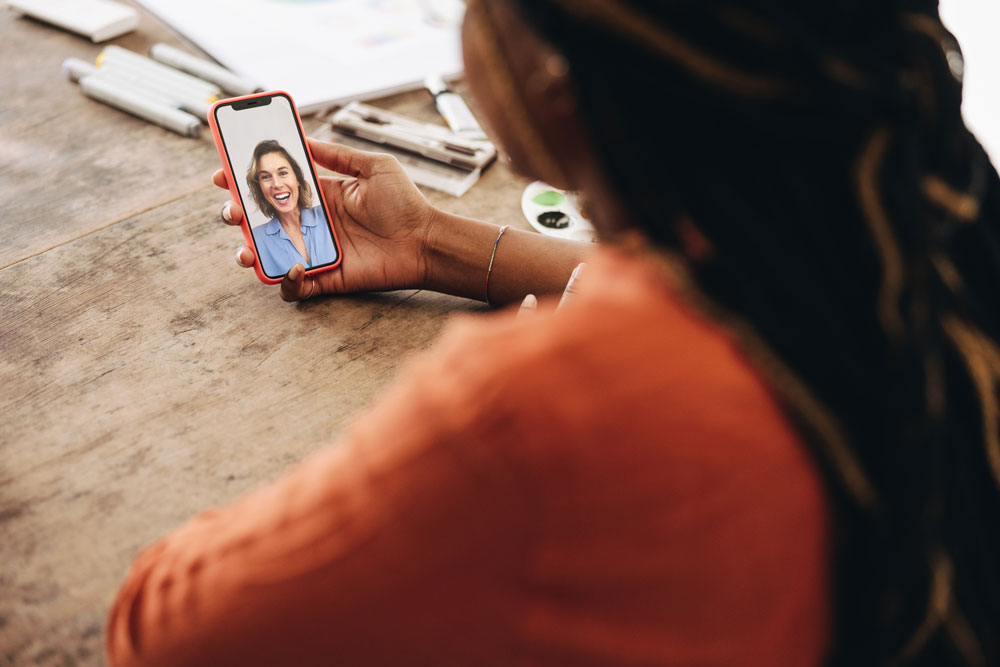 A person holding a smartphone is video calling someone who is smiling. The caller sits at a wooden table with papers and pens scattered nearby. Only the callers back is visible, while the friends face is on the screen.