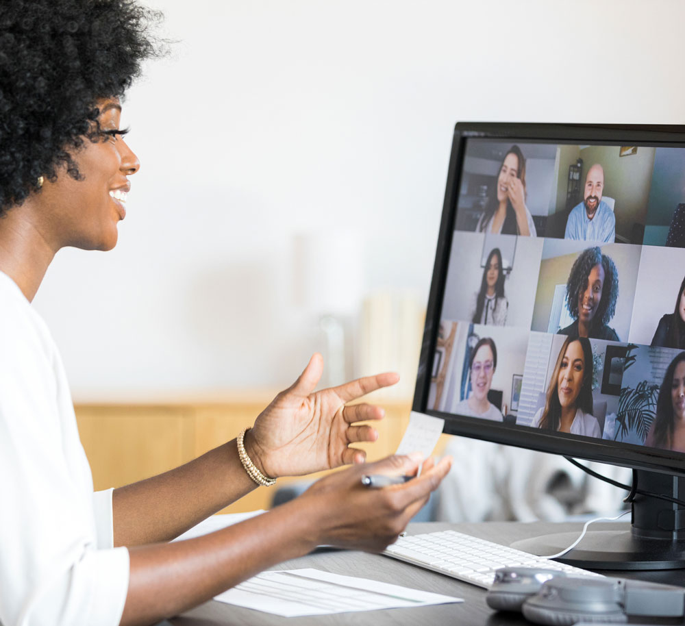 A woman sits at a desk, participating in a video call with nine people displayed on her computer monitor, gesturing with her hand and holding a pen.