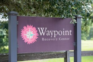 A sign for Waypoint Recovery Center stands outdoors, featuring a compass design and surrounded by greenery.