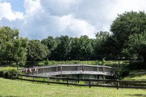 A wooden footbridge crosses over a small pond surrounded by trees and greenery under a partly cloudy sky.