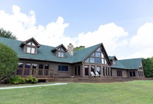 A large log cabin house with green roof, several dormer windows, a stone chimney, and a spacious wooden deck, set on a grassy lawn under a partly cloudy sky.