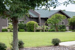 A wooden bench swing stands in a green yard with manicured bushes and trees in front of a brown house with a green roof.