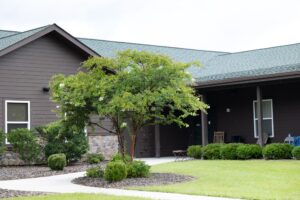 A single-story brown house with a covered porch, green lawn, shrubs, a small tree, and a stone accent on the exterior wall.