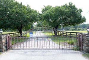 A closed metal gate blocks a driveway leading to a rural property with green grass, large trees, and wooden fencing. Multiple security and warning signs are posted on the gate.